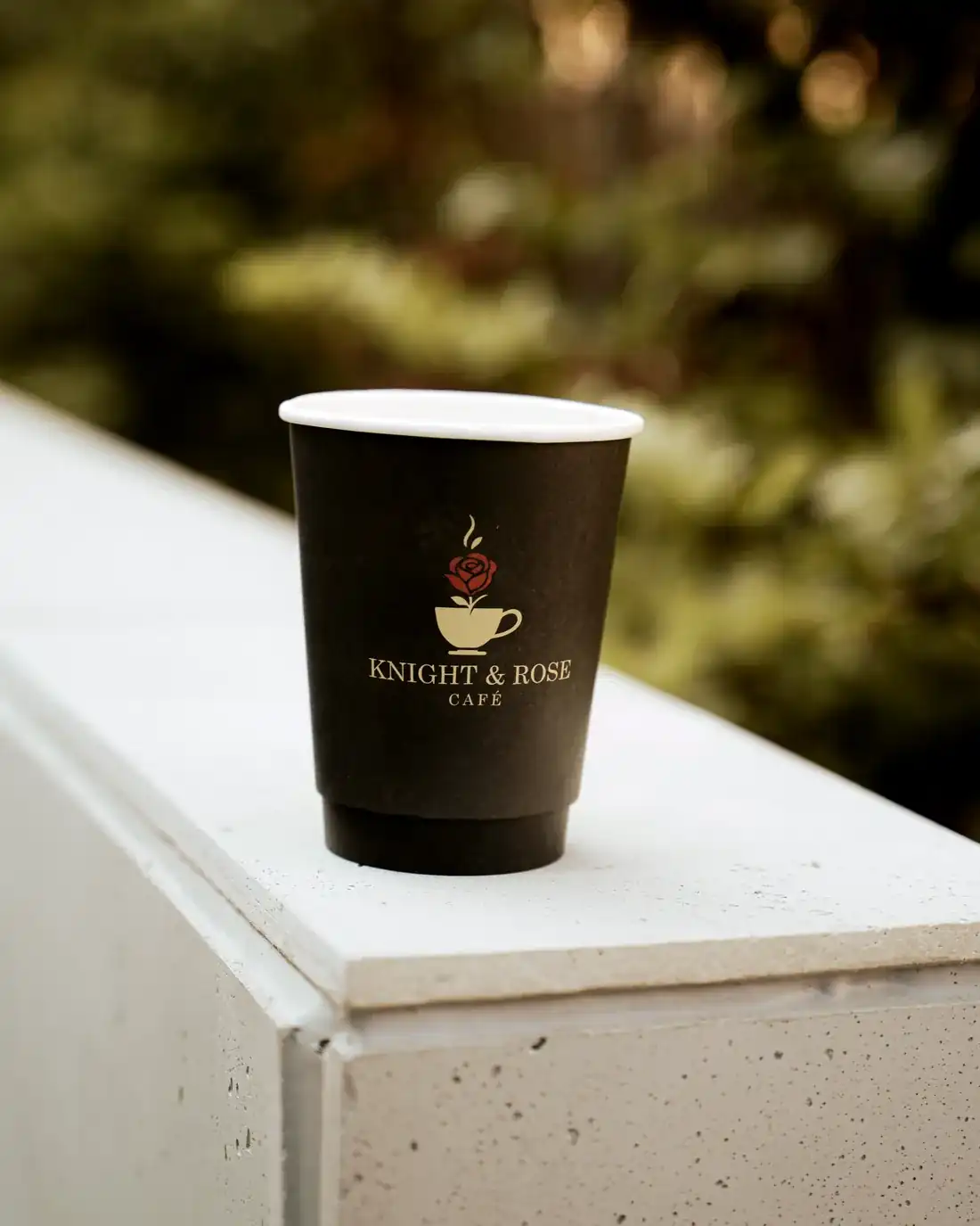 A Knight & Rose Café cup with its gold rose emblem, resting on a stone ledge in afternoon light.